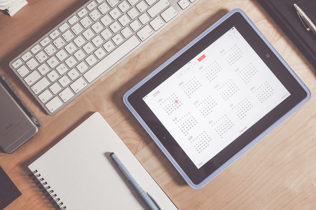 ipad with calendar open, next to a pen and pad of paper, a keyboard and an iphone on a desk